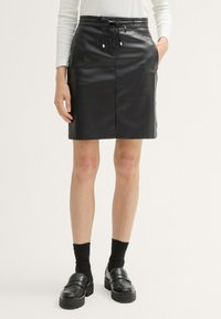 Schwarzer Kunstlederrock mit einer Zugbandtaillierung, seitlichen Taschen und einer glatten Textur, der über dem Knie endet. Getragen mit schwarzen Slippern und Socken.