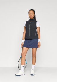 Svart quiltet vest med ståkrage, hvit kortermet skjorte, marineblå tennisrugh, hvite sko, holder en tenniskølle, med håndleddbånd.
