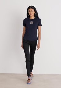 Marineblå kortermet skjorte med et rundt grafisk logo på brystet, sammen med svarte skinny jeans og blå sandaler med stropper.