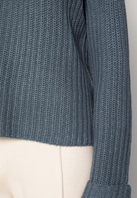 Närbild av ribbat, stickat mörkgrått tröjärm och nederkant, parat med ljusbeiga byxor.