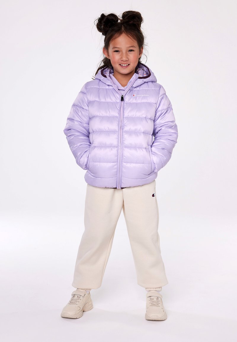 Giacca a vento viola con cuciture orizzontali, zip frontale e cappuccio. Accoppiata con pantaloni della tuta color crema e scarpe da ginnastica chiare.