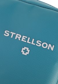 Teal stof med prægede teksten "STRELLSON" i sølv, sammen med et lille kors emblem, på en struktureret baggrund.