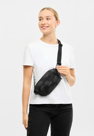 Borsa a tracolla nera con superficie testurizzata, tracolla regolabile e chiusura con zip, indossata sopra una maglietta bianca e jeans neri.