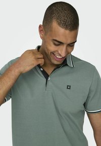 Grünes Polohemd aus Baumwolle. Mit schwarzem Kragen mit weißen Streifen, Knopfleiste und einem dezenten Logo auf der Brust.