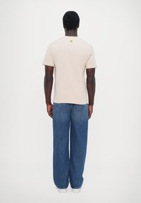 Camiseta de manga corta color beige claro con un pequeño logo sonriente verde en la espalda, combinada con jeans azules holgados y zapatos blancos.