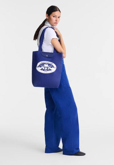 Blue canvas tote bag with a front logo patch. The bag features a top snap closure and sturdy handles. Accompanied by a white shirt and scarf.