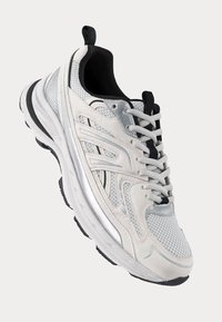 Chaussure de sport avec une tige en mesh blanche, des accents argentés et des détails noirs. Elle présente un col rembourré, une fermeture à lacets et une semelle texturée.