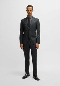Completo grigio scuro con vestibilità su misura, giacca monopetto e pantaloni abbinati. Tessuto liscio, cravatta nera e scarpe nere lucide.