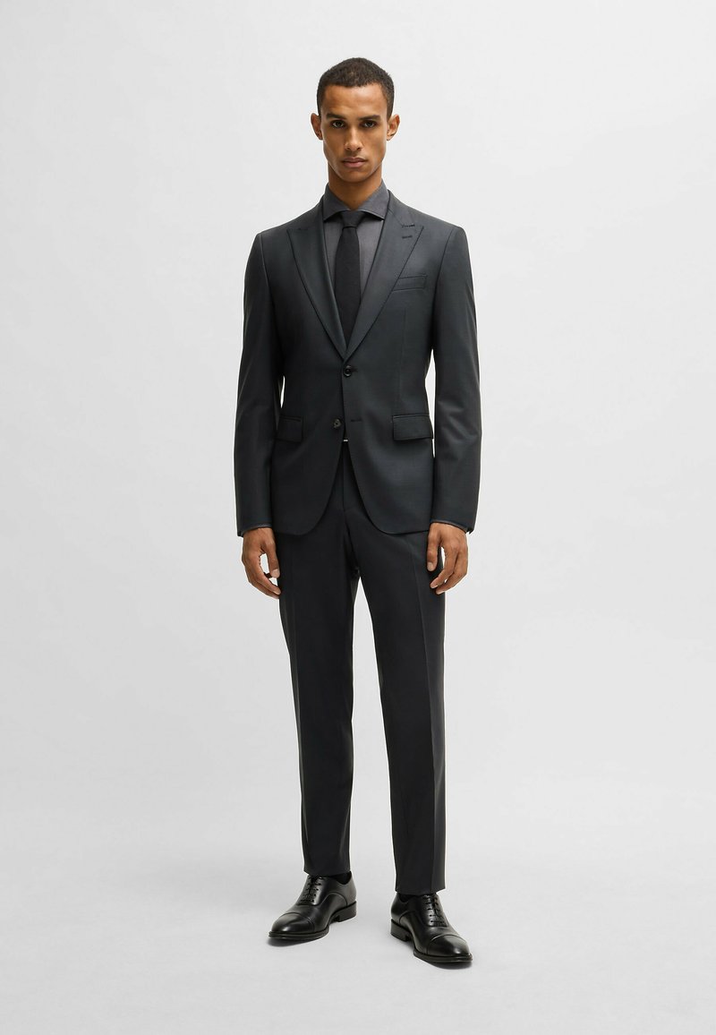 Completo grigio scuro con vestibilità su misura, giacca monopetto e pantaloni abbinati. Tessuto liscio, cravatta nera e scarpe nere lucide.