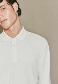Balta ilgomis rankovėmis polo marškinėliai, pasižymintys tekstūruotu audiniu, minkštu apykakliu ir trimis mygtukais; minimalistinis dizainas su subtiliomis detalėmis.