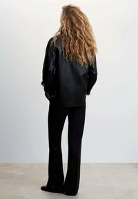 Musta nahkatakki rentoa siluettia, yhdistettynä muotoiltuihin mustiin housuihin. Teksturoitu tukka laskeutuu hartioille. Minimalistinen tausta.