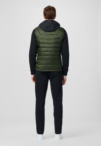 Zielona kamizelka puchowa z poziomym przeszyciem, noszona na czarnej bluzie z kapturem, w zestawie z czarnymi spodniami i białymi sneakersami. Minimalistyczny design.