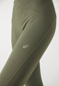 Olivgrüne Leggings aus glattem, dehnbarem Stoff. Mit flachen Nähten und einem kleinen weißen Logo auf dem Oberschenkel. Hohe Taille.
