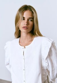 Blusa blanca de botones con mangas acampanadas, hecha de tela ligera. Presenta sutiles detalles bordados y un escote redondo.