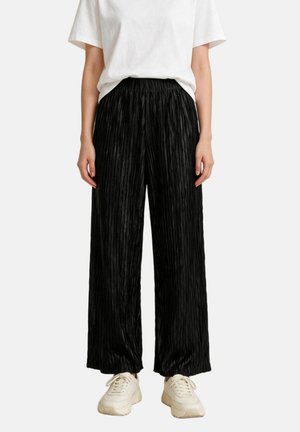 Personne portant un pantalon noir texturé à jambes larges, une chemise blanche à manches courtes et des baskets blanches sur un fond uni.