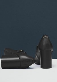 Pár černých kožených bot na blokovém podpatku s přezkami, jedna bota je vztyčená a druhá leží na boku na tmavě modrém pozadí.