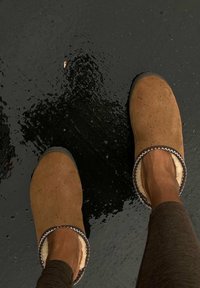 Bruine suède klompen met een witte fleece voering en decoratieve stiksels, geplaatst tegen een natte zwarte ondergrond. Ronde neus en instapontwerp.