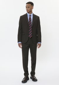 Brązowy garnitur o dopasowanym kroju, z dwoma guzikami i jedną kieszenią na piersi. W zestawie z jasnoniebieską koszulą i wąskim krawatem w paski. Błyszczące czarne buty.