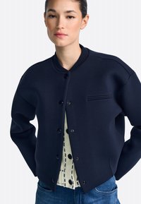 Donkerblauwe bomberjack gemaakt van gestructureerde stof, met een geribde kraag, knoopsluiting en een kleine horizontale streep op de borst.