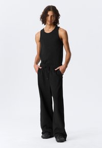 Νεαρό άτομο με κατσαρά μαλλιά στέκεται με τα χέρια στις τσέπες, φορώντας μαύρο αμάνικο τοπ, φαρδιά μαύρα παντελόνια και μαύρα παπούτσια.