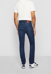 Mörkblå denimjeans med en figurnära design, bakfickor och en brun läderlapp vid midjan. I kombination med en enfärgad vit T-shirt och vita sneakers.