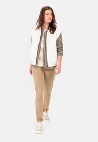 Hvid fleece vest over en grøn bomberjakke, stribet skjorte, khaki bukser og grå sneakers. Modellen står foran en neutral baggrund.