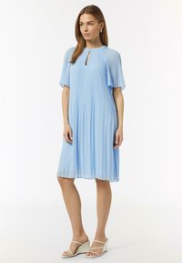 Himmelblaues Faltenkleid mit kurzen Ärmeln und einem Schlüsselloch-Ausschnitt. Der Stoff wirkt fließend und weich. Tragen mit weißen Keilsandaletten.