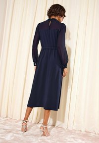 Vestido azul marino con mangas largas translúcidas, cintura elástica y puños con estampado floral. Presenta un escote en forma de llave en la parte posterior y una falda de longitud media, combinado con tacones.