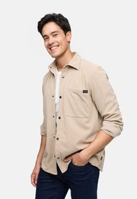 Camisa de pana beige con cierre de botones en la parte delantera, un bolsillo en el pecho y mangas remangadas, combinada con jeans azul oscuro.