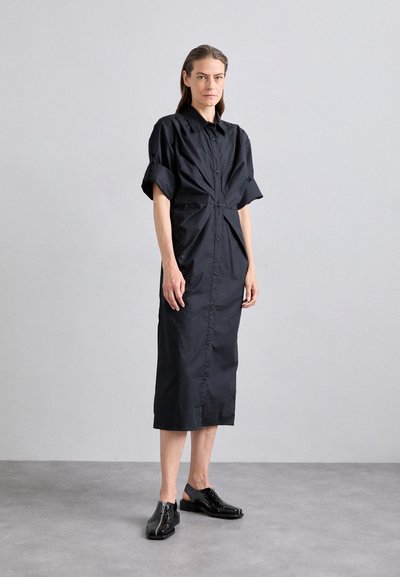 Zwarte jurk met korte opgerolde mouwen, knopen aan de voorkant en een gathered taille. Textuurstof met een knie-lange zoom. Paar zwarte schoenen.