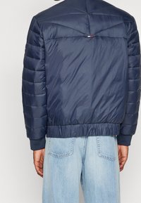 Marinblå puffjacka med quiltat mönster, elastiska ärmar och nederkant, utformad med en slät texturfinish. Baksidan har en dekorativ söm.