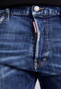 Donkerblauwe spijkerbroek met een vervaagde afwerking, voorzien van koperen stiksels en een zichtbaar merketiket. Knoopsluiting bij de taille.