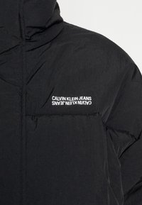 Svart puffjacka med hög krage, med synliga sömmar, en framficka och vit "Calvin Klein Jeans"-logotyp på bröstet.