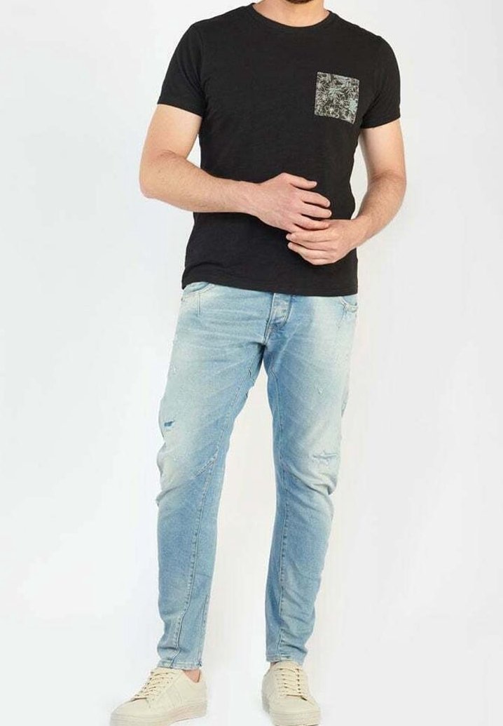 Mand iført sort t-shirt med mønstret brystlomme, lyseblå slidte jeans og off-white sneakers, stående mod en hvid baggrund.