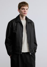 Μαύρο ελαφρύ μπουφάν με υφή, κλείσιμο με φερμουάρ και μεγάλο γιακά. Φορεμένο πάνω από ανοιχτόχρωμο μπλουζάκι με στρογγυλή λαιμόκοψη.