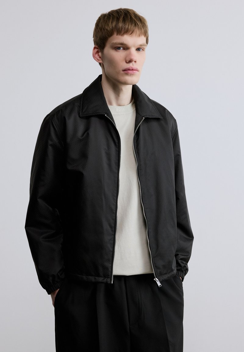 Μαύρο ελαφρύ μπουφάν με υφή, κλείσιμο με φερμουάρ και μεγάλο γιακά. Φορεμένο πάνω από ανοιχτόχρωμο μπλουζάκι με στρογγυλή λαιμόκοψη.