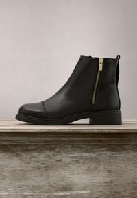 Bottine en cuir noir avec fermeture éclair latérale et petit talon, posée sur une surface en bois sur un fond uni.