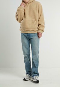 Sudadera con capucha de forro polar beige con un bolsillo frontal, combinada con jeans de mezclilla azul claro y zapatillas plateadas. Textura suave y corte relajado.