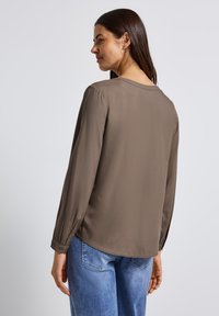 Blouse marron à manches longues avec un col rond, texture douce et poignets boutonnés, associée à un jean bleu clair dans un cadre décontracté.