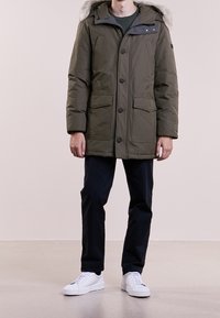 Olivekowa zielona parka z kapturem wyłożonym futrem, zapinana na guziki, z dwoma przednimi kieszeniami i teksturowanym wykończeniem. Noszona z ciemnymi spodniami i białymi trampkami.