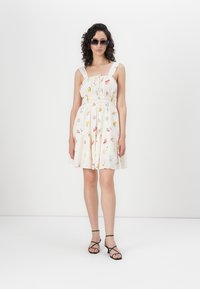 Romantische jurk met bloemenpatroon, crème kleur, voorzien van een smocktail en brede bandjes, met een gelaagde rok. Gepaarde met zwarte sandaaltjes met straps en een zonnebril.