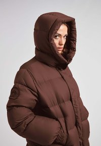 Bruine pufferjas met een capuchon, met een gequilt ontwerp, brede mouwen en een ronde logopatch op de schouder. Zachte, glanzende textuur.