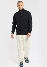 Pull en maille marine avec un col haut, doté de quatre boutons. Associé à un pantalon en velours côtelé écru et des baskets bleues.