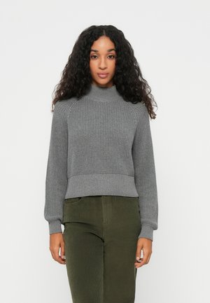 Grå ribbestrikket genser med høy hals og lange ermer, sammen med mørkegrønne kordfløyelsbukser. Stoffet virker teksturert og varmt.