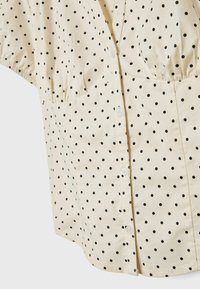 Beige button-up shirt with black polka dots, featuring a gathered waist, short sleeves, and a classic collar. White buttons accent the front.