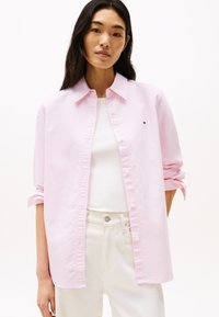 Camicia button-up a righe rosa chiaro con colletto classico, dotata di maniche arrotolabili. Indossata sopra un top bianco e abbinata a pantaloni chiari.