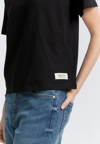 Zwarte korte mouwen T-shirt van katoen, met een recht model en een gestikt label "DENIM TOM TAILOR" bij de zoom. Gemaakt in combinatie met blauwe spijkerbroek.