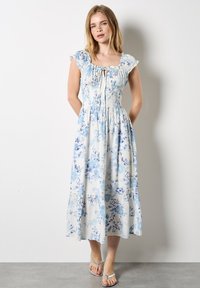 Robe entièrement florale en bleu clair et blanc, avec un corsage smocké, des manches volantées et une jupe à volants. Le tissu semble léger et fluide.