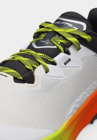 Αθλητικό παπούτσι με λευκό επάνω μέρος από δίχτυ, μαύρα και νέον κίτρινα κορδόνια, μαύρη γλώσσα με την ένδειξη "ALTRA TIMP 6" και σόλα με πορτοκαλί-κίτρινο διαβάθμιση.