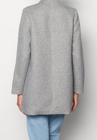 Abrigo de lana gris texturizado con corte recto, mangas largas, cuello con muesca y detalle de costura en la parte trasera; combinado con vaqueros de mezclilla azul claro.